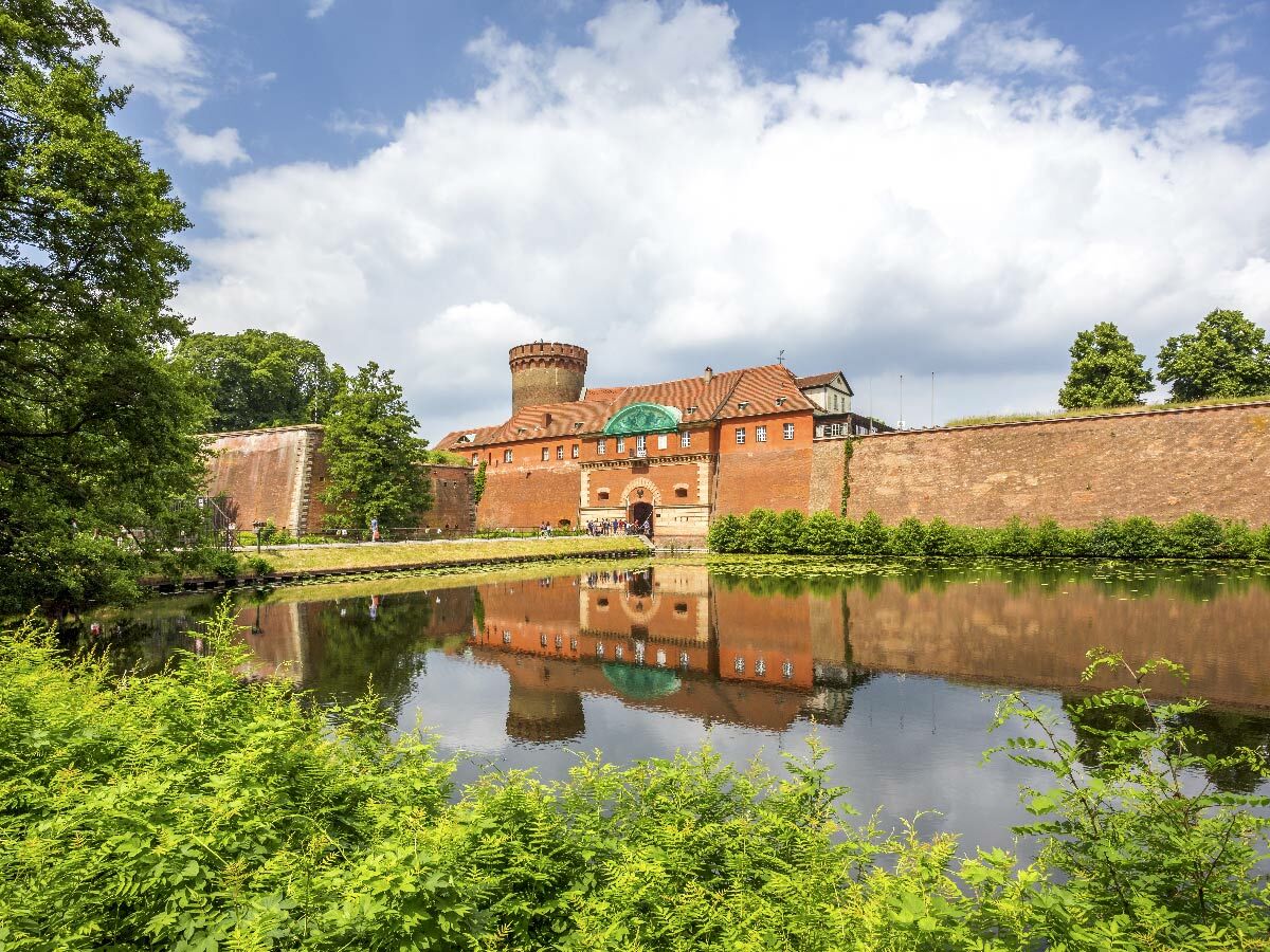 Rote Backsteinfestung mit Turm und Wassergraben, umgeben von grünen Bäumen unter bewölktem Himmel.