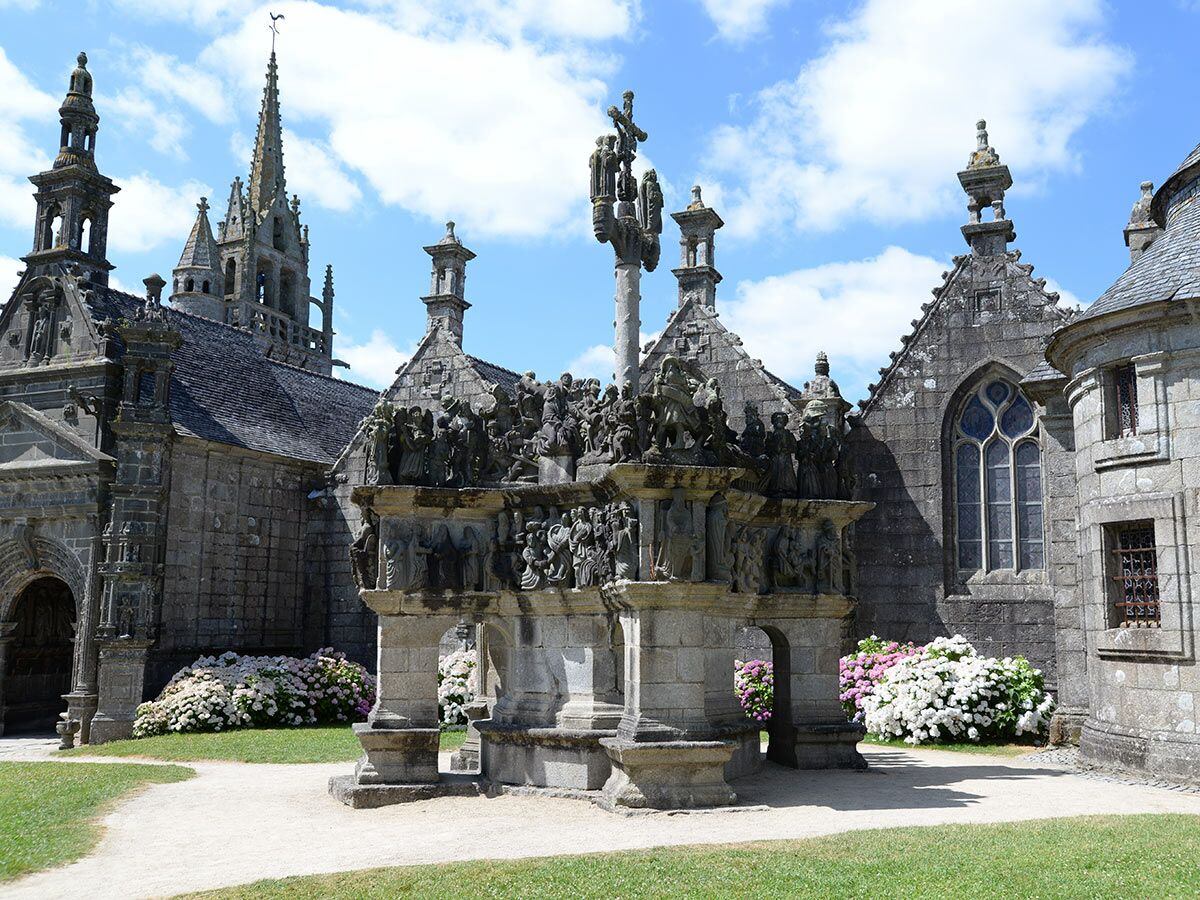 Historischer Kalvarienberg und Kirche aus Stein in Saint-Thégonnec, Bretagne, mit blühenden Hortensien unter blauem Himmel.