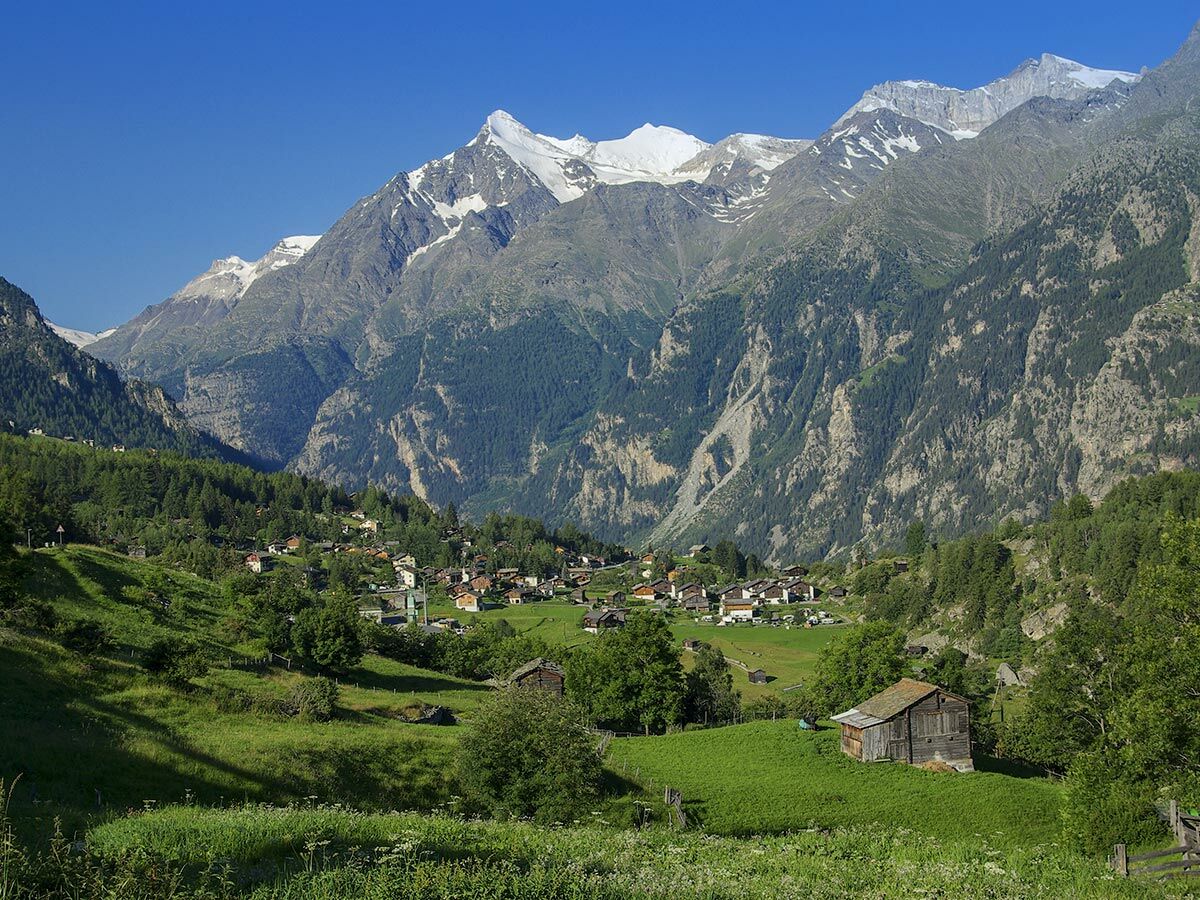 Panorama einer idyllischen Alpenlandschaft mit einem kleinen Bergdorf im Tal, umgeben von grünen Hügeln und majestätischen schneebedeckten Gipfeln unter blauem Himmel.