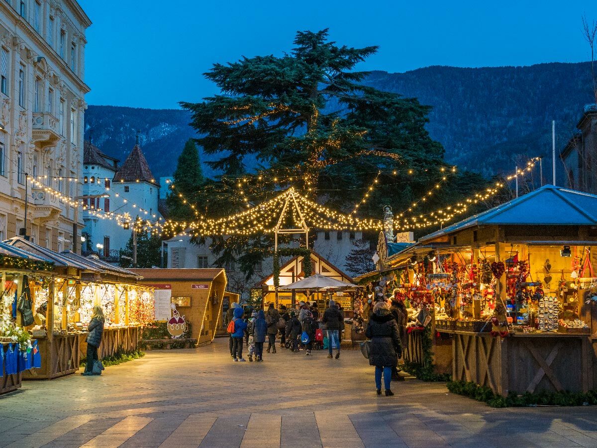 Festlicher Weihnachtsmarkt in Meran bei Dämmerung mit hell beleuchteten Ständen, Besuchern und einem historischen Gebäude im Hintergrund.