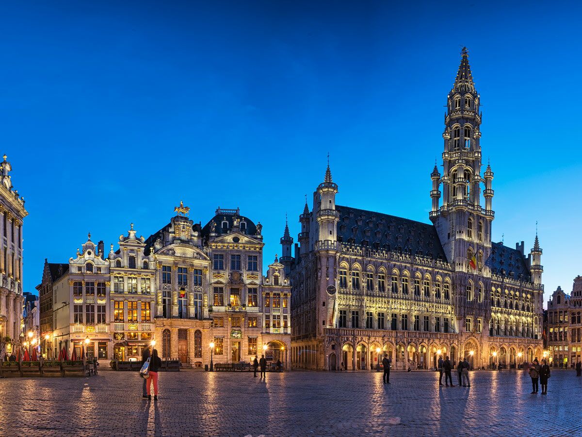 Historischer Grand Place in Brüssel bei Abend, mit beleuchtetem Rathaus, opulenten Gildehäusern und Menschen auf dem Platz.