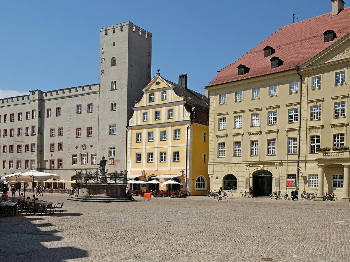Blick auf den belebten Straubinger Stadtplatz mit Stadtturm, historischen Gebäuden, Brunnen und Café-Terrassen an einem sonnigen Tag.