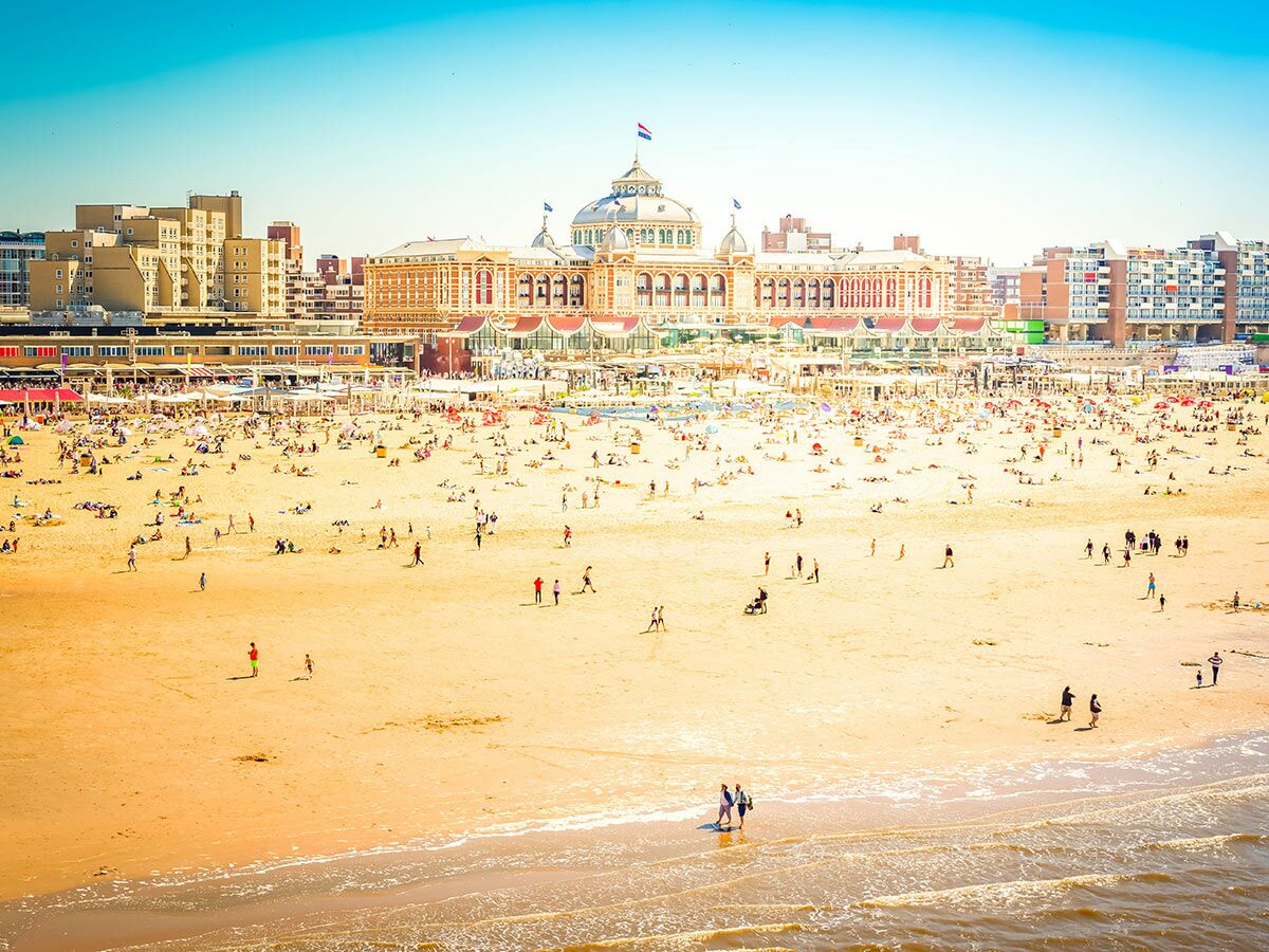 Belebter Strand von Scheveningen mit dem ikonischen Kurhaus und vielen Menschen, die das sonnige Wetter genießen.