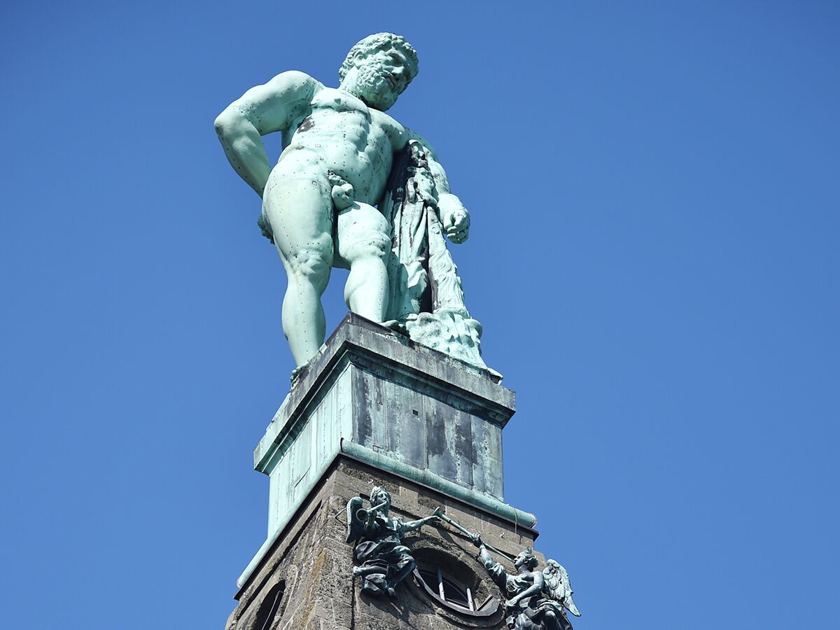 Majestätische Bronzestatue eines muskulösen Mannes, vermutlich Herkules, mit grüner Patina auf Steinsockel vor blauem Himmel.