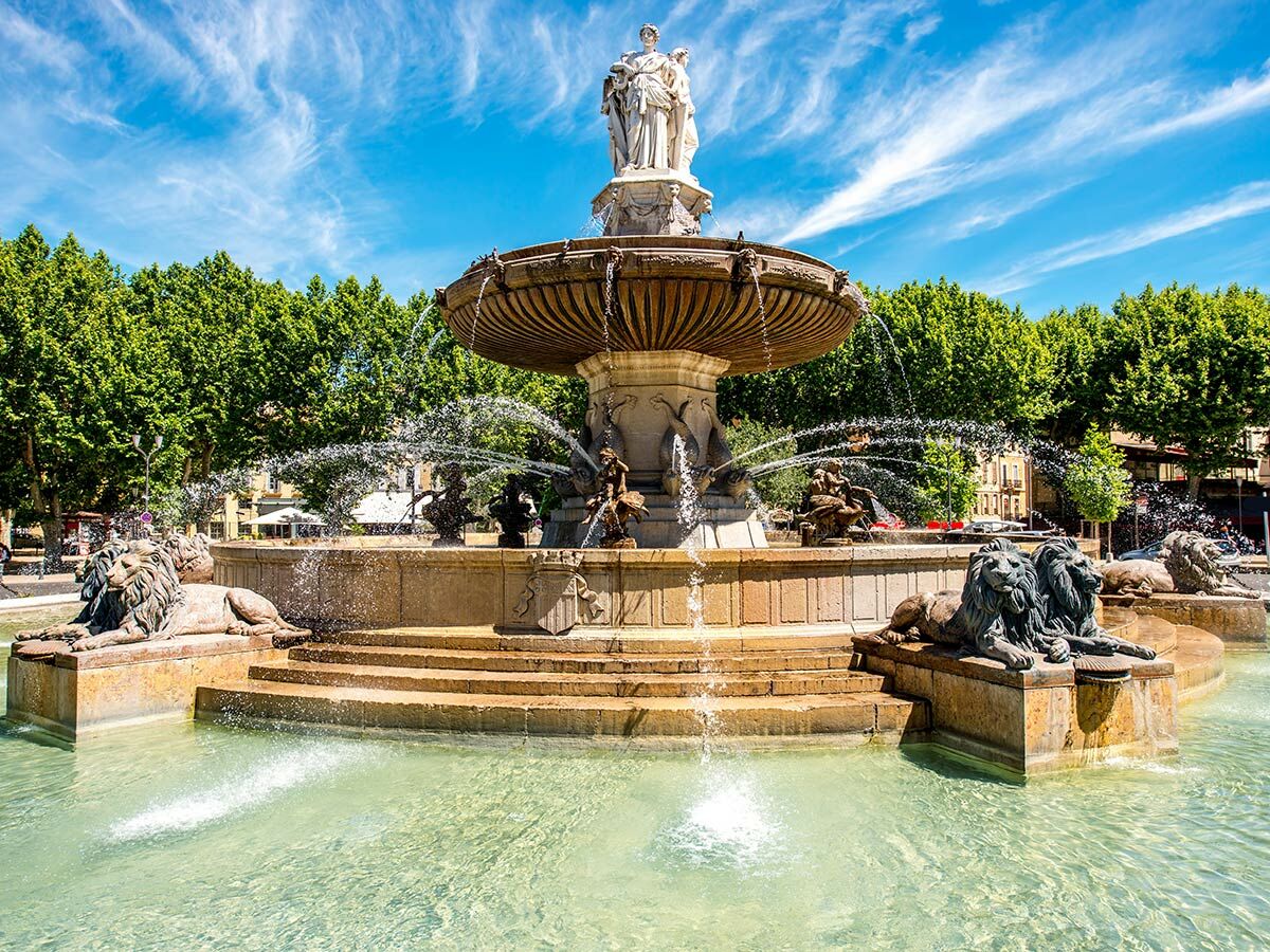 Großer, historischer Brunnen mit Löwenstatuen, Frauenstatue und sprudelndem Wasser unter blauem Himmel in Aix-en-Provence.