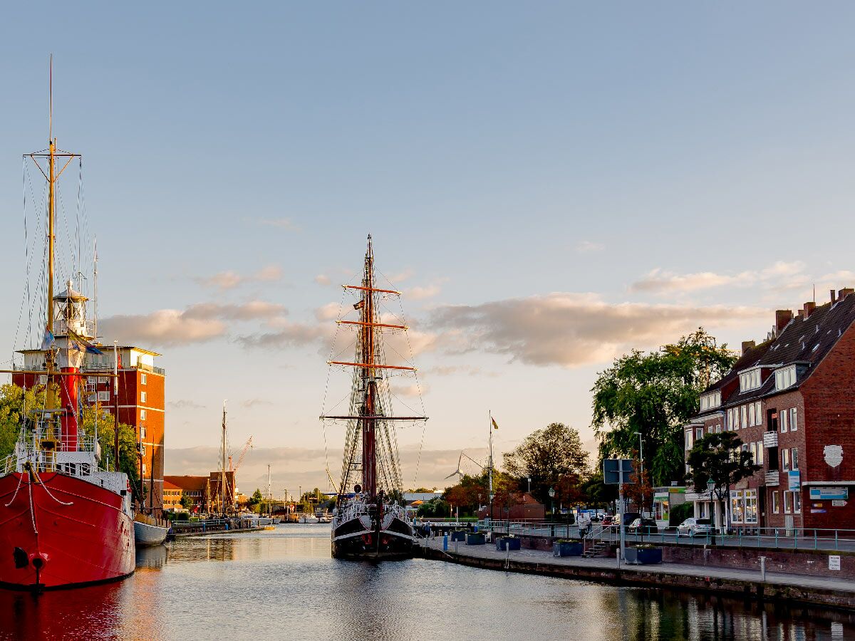 Zwei historische Schiffe in einem Kanal, darunter ein rotes Feuerschiff, in einem Hafen mit alten Backsteingebäuden bei Abendlicht.
