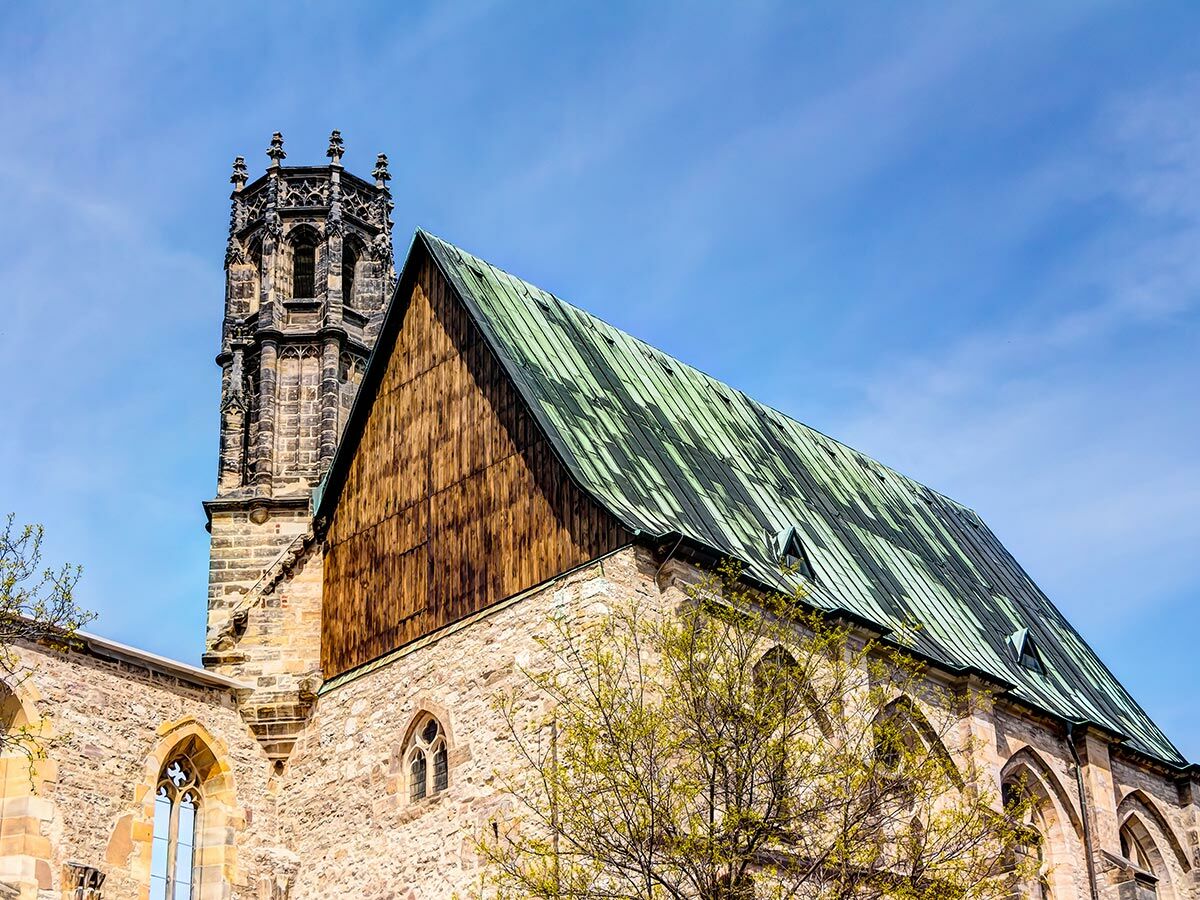 Historische gotische Kirche mit hohem Turm, steinernen Mauern, Holzfassade und grünem Metalldach unter blauem Himmel im Frühling.
