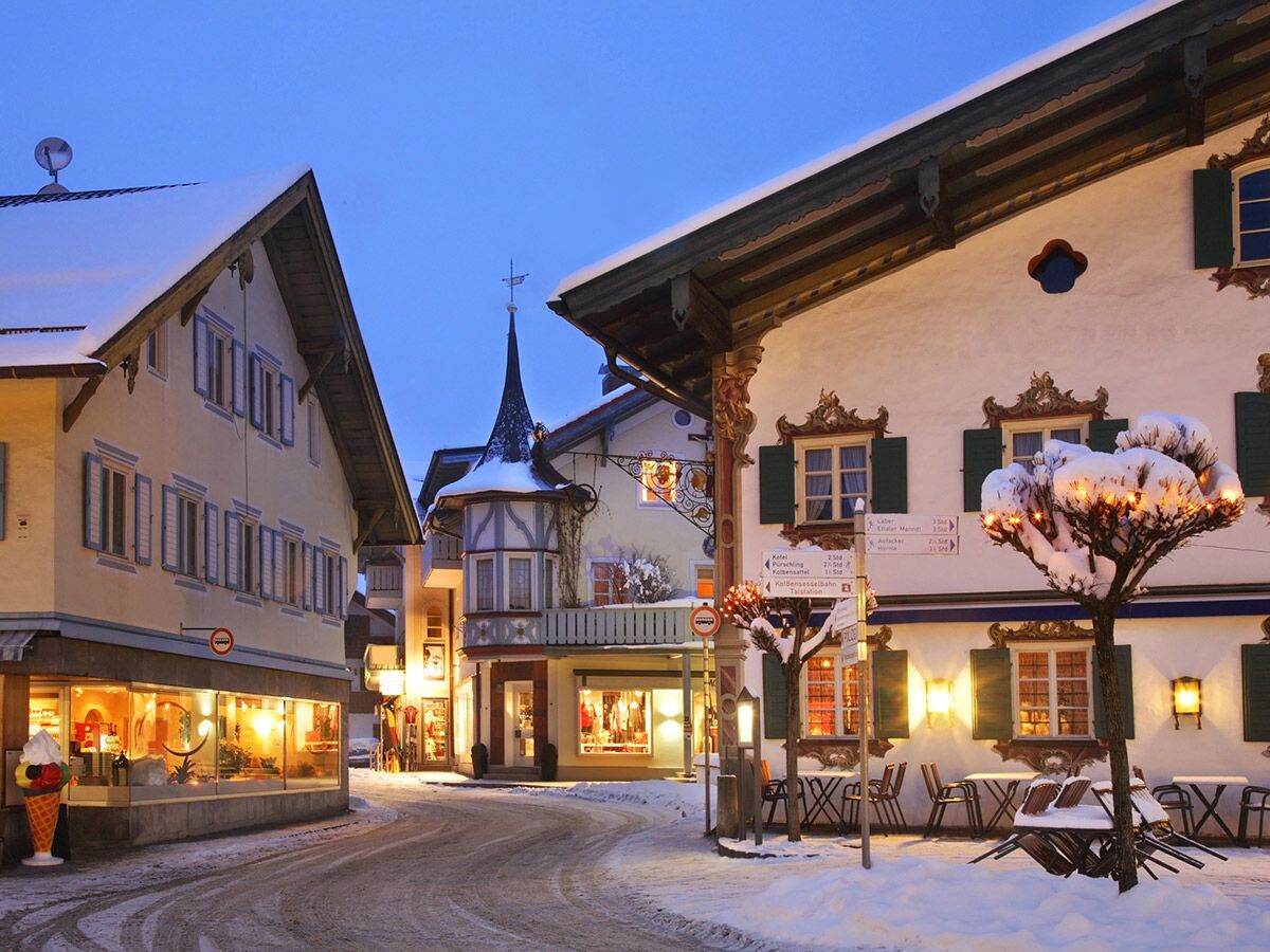 Schneebedeckte Straße in bayerischem Winterdorf bei Dämmerung, beleuchtete Gebäude und festliche Lichter an Bäumen. (124 Zeichen)