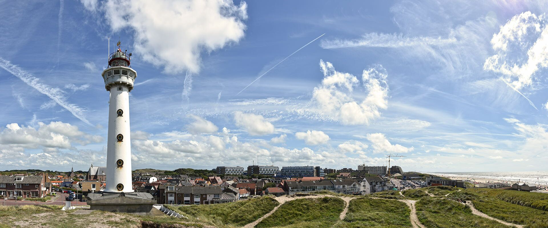 Panoramablick auf einen weißen Leuchtturm, eine Küstenstadt, grüne Dünen und einen belebten Sandstrand mit Meer unter blauem Himmel.