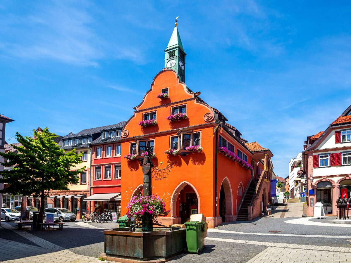 Leuchtend orangefarbenes Rathaus mit grünem Uhrturm und Blumenschmuck auf einem sonnigen Marktplatz in einer europäischen Altstadt.