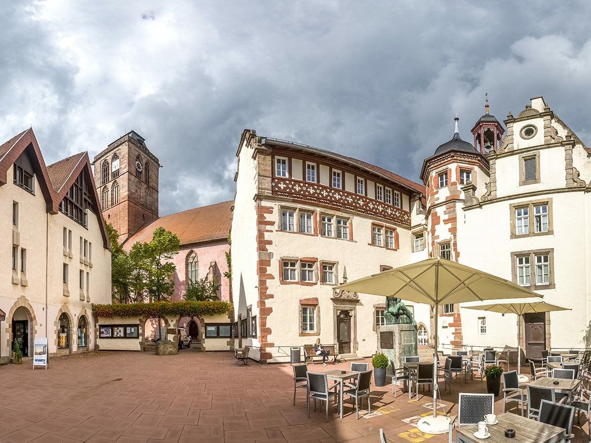 Blick auf den historischen Klosterhof Maulbronn mit Abteikirche und Café, Menschen entspannen im Freien an einem bewölkten Tag.