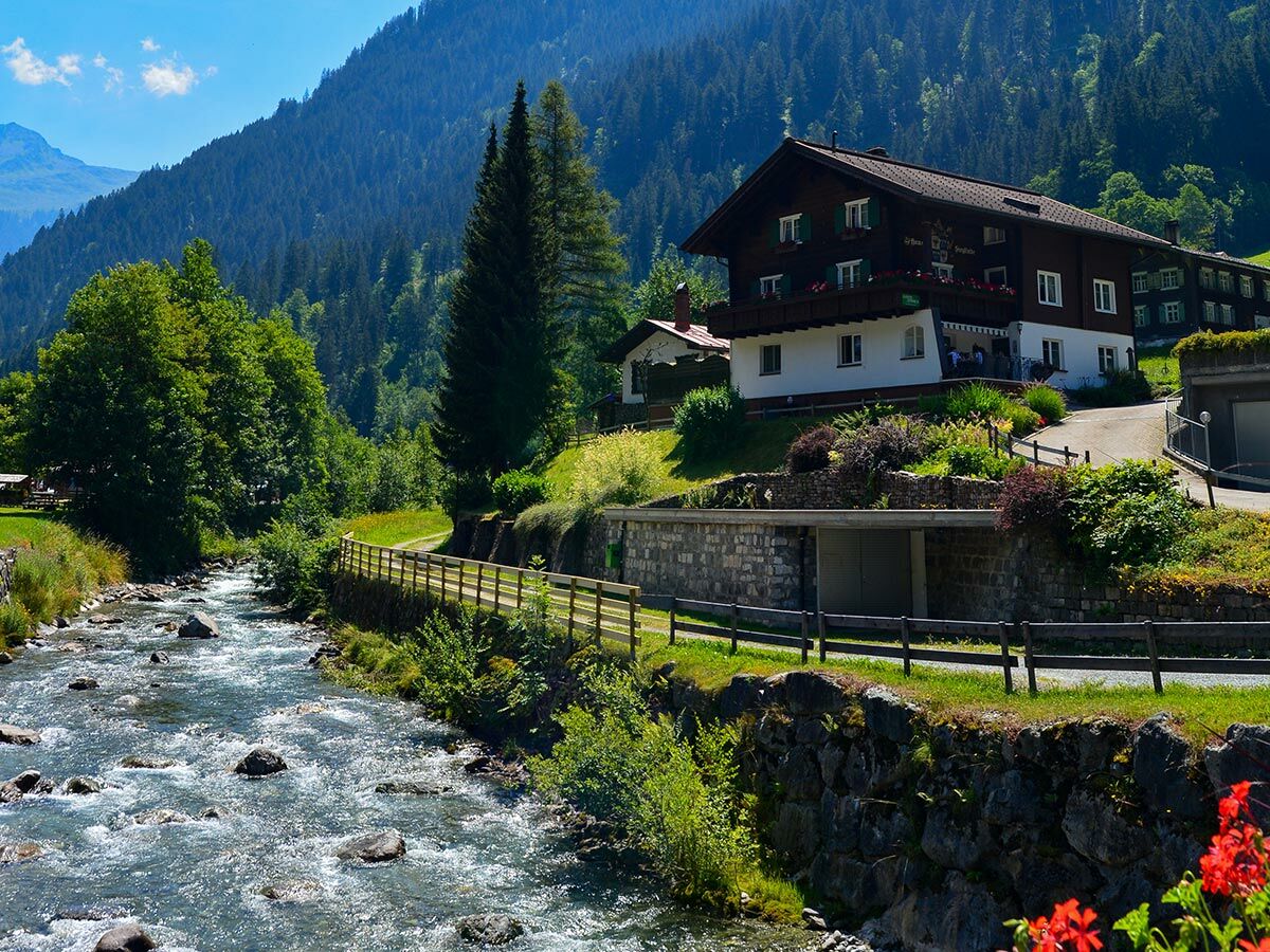 Traditionelles Alpenhaus mit grünen Fensterläden und Balkonen am rauschenden Bergbach, umgeben von Wäldern und Bergen.