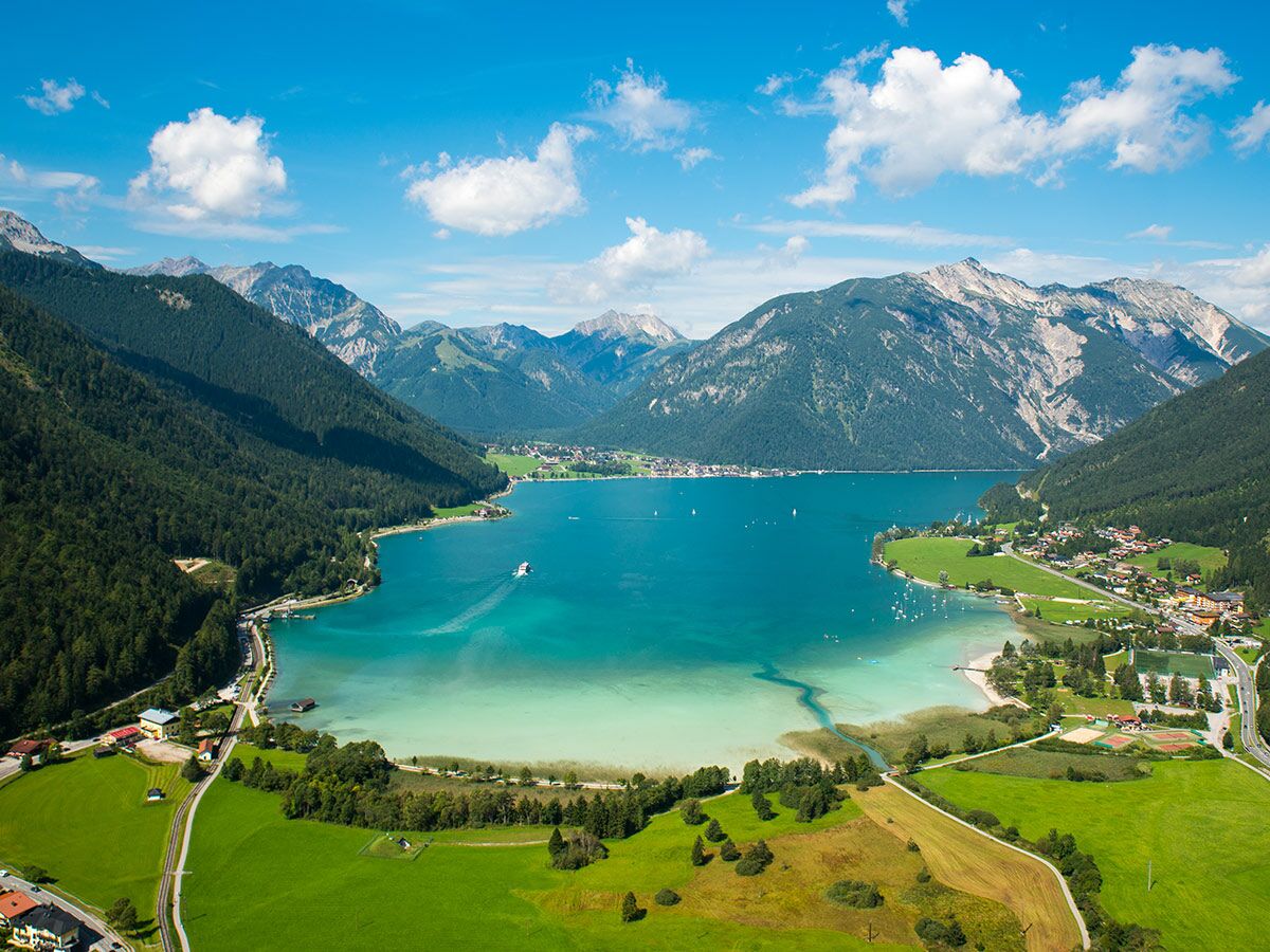 Azurblauer Achensee in Tirol, umgeben von grünen Wäldern und majestätischen Alpen. Boote auf dem Wasser, idyllische Dörfer am Ufer.