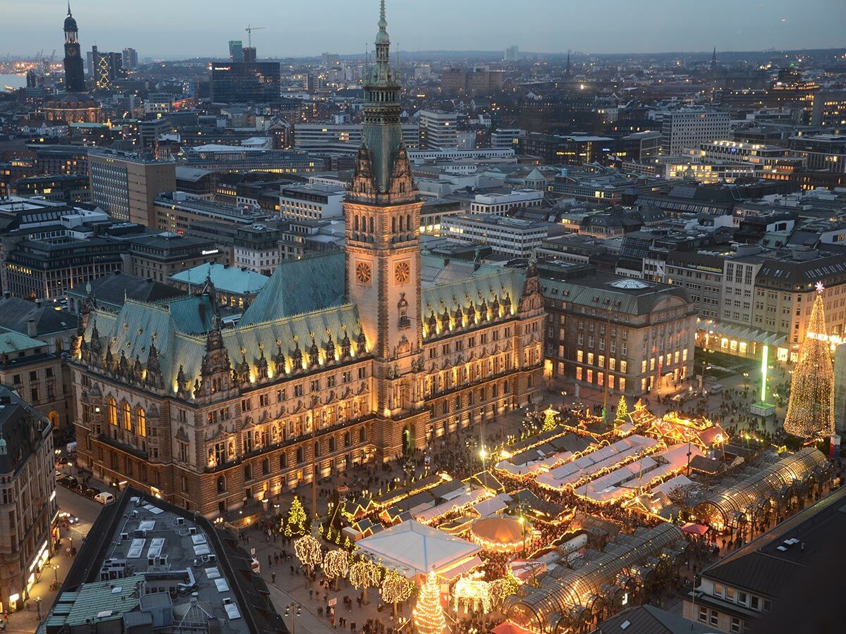 Luftbild des beleuchteten Hamburger Rathauses bei Dämmerung mit einem geschäftigen Weihnachtsmarkt voller Lichter und Menschen davor.