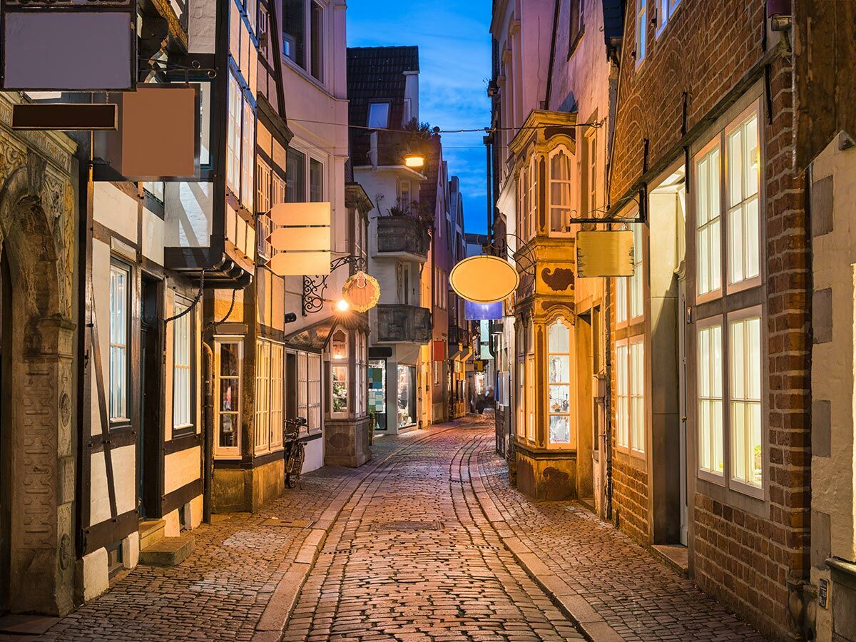 Schmale historische Gasse mit Kopfsteinpflaster und alten Fachwerkhäusern bei Abenddämmerung, beleuchtete Fenster und Schilder.