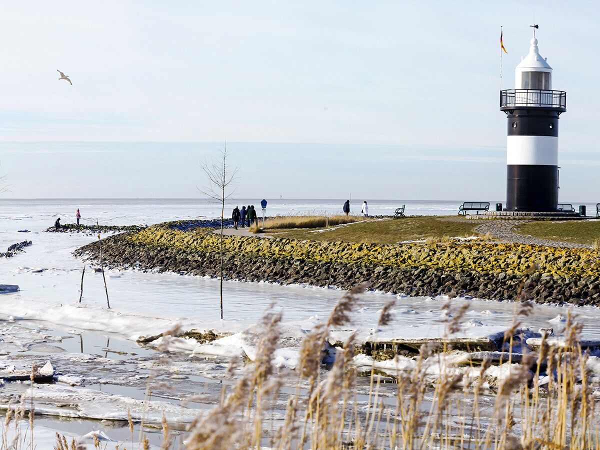 Schwarz-weißer Leuchtturm an vereister Küste mit Menschen, Möwe und Eisschollen an sonnigem Wintertag in Deutschland.