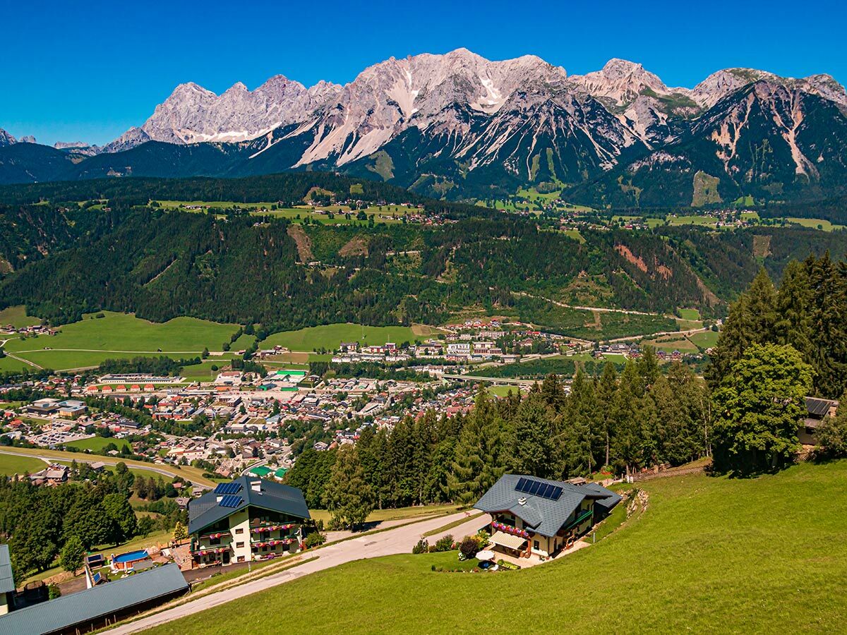Panoramablick über eine grüne Tal- und Berglandschaft mit einer Stadt, dichten Wäldern und sonnenbeschienenen Alpen, im Vordergrund zwei Häuser mit Solarpaneelen.