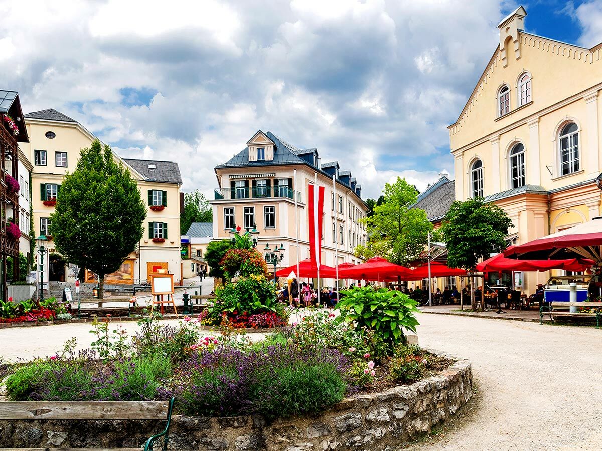 Belebter österreichischer Dorfplatz mit historischen Gebäuden, blühenden Blumenbeeten, Cafés unter roten Sonnenschirmen.