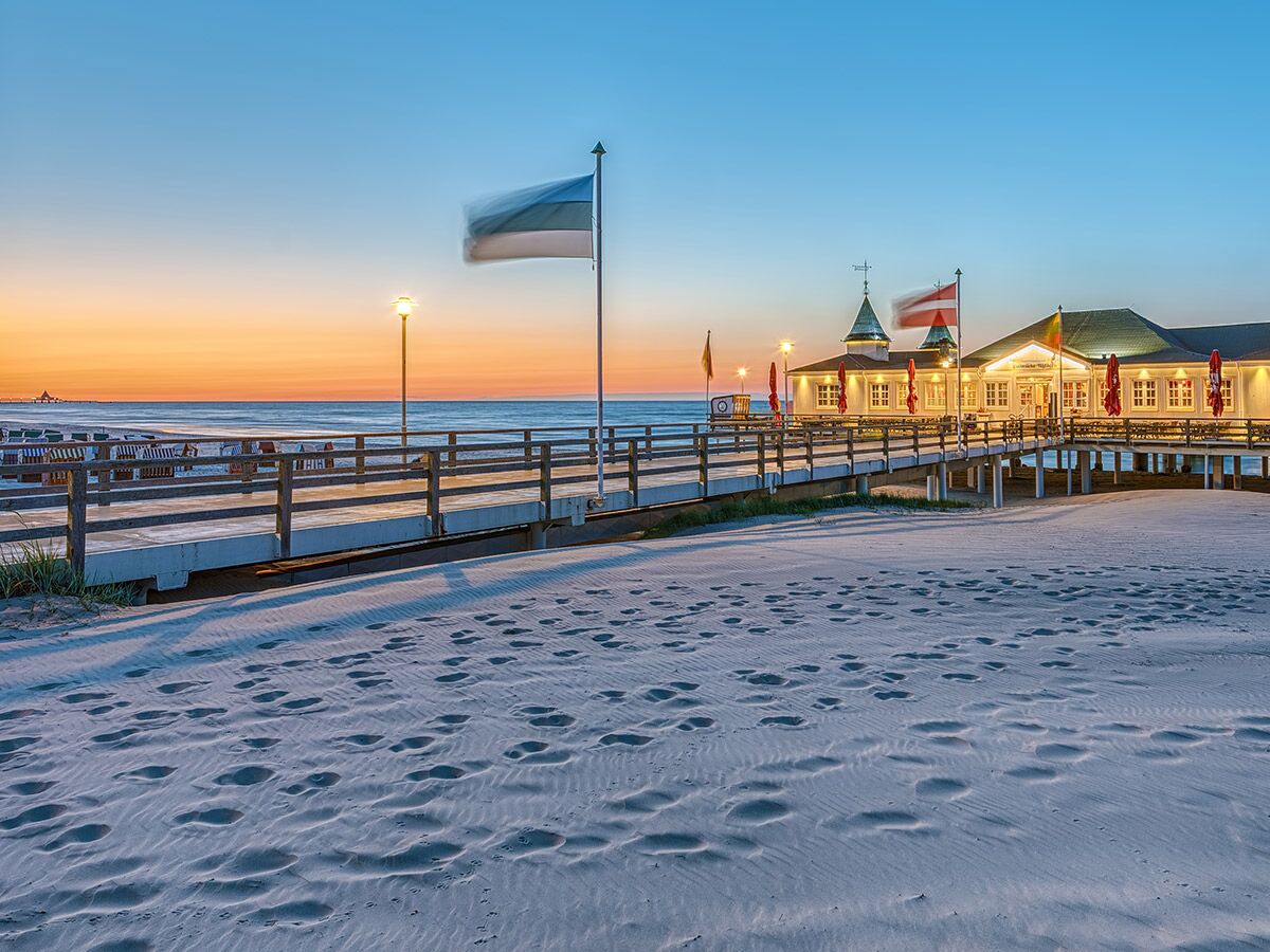 Abendlicher Blick auf beleuchtete Seebrücke mit Restaurant am Sandstrand und Fußspuren bei Sonnenuntergang über der Ostsee.