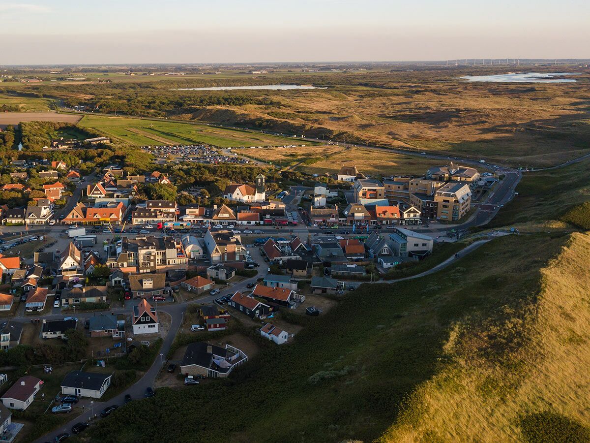 Luftaufnahme einer idyllischen Küstenstadt mit vielen Häusern, Straßen und Autos in einer weiten Dünenlandschaft, aufgenommen im goldenen Abendlicht.