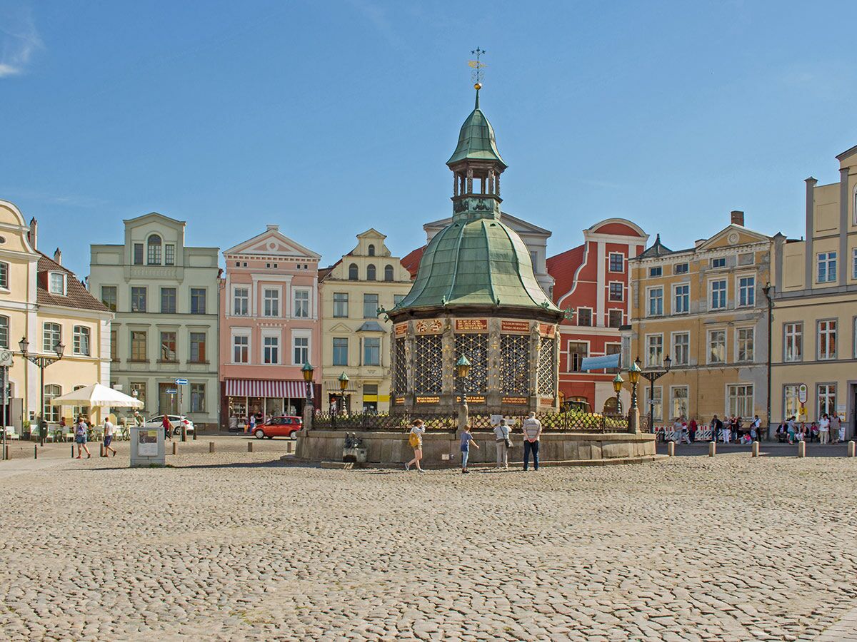 Sonnenbeschienener Marktplatz in Wismar mit Wasserkunst-Brunnen, historischen Giebelhäusern und flanierenden Personen.