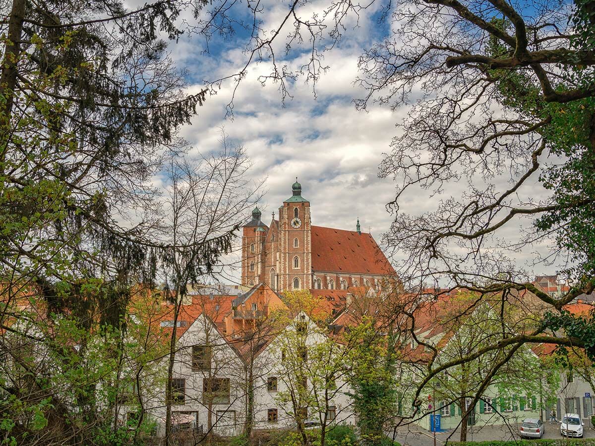 Panoramablick auf die Stiftskirche St. Martin in Landshut, Bayern, mit imposanten Backsteintürmen, roten Dächern und grünen Bäumen unter bewölktem Himmel.