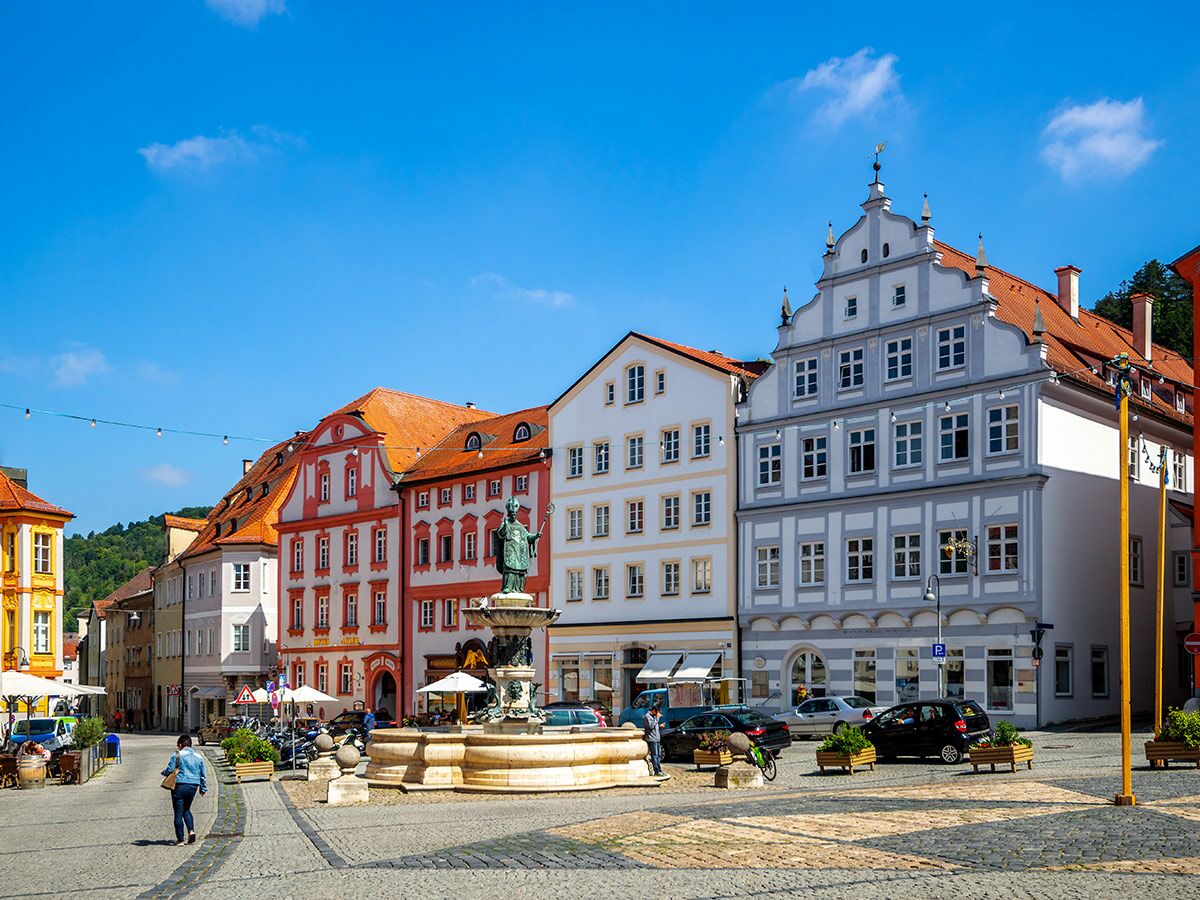 Blick auf einen sonnigen historischen Marktplatz mit bunten Giebelhäusern, einem Brunnen mit Statue, Cafés und Menschen.