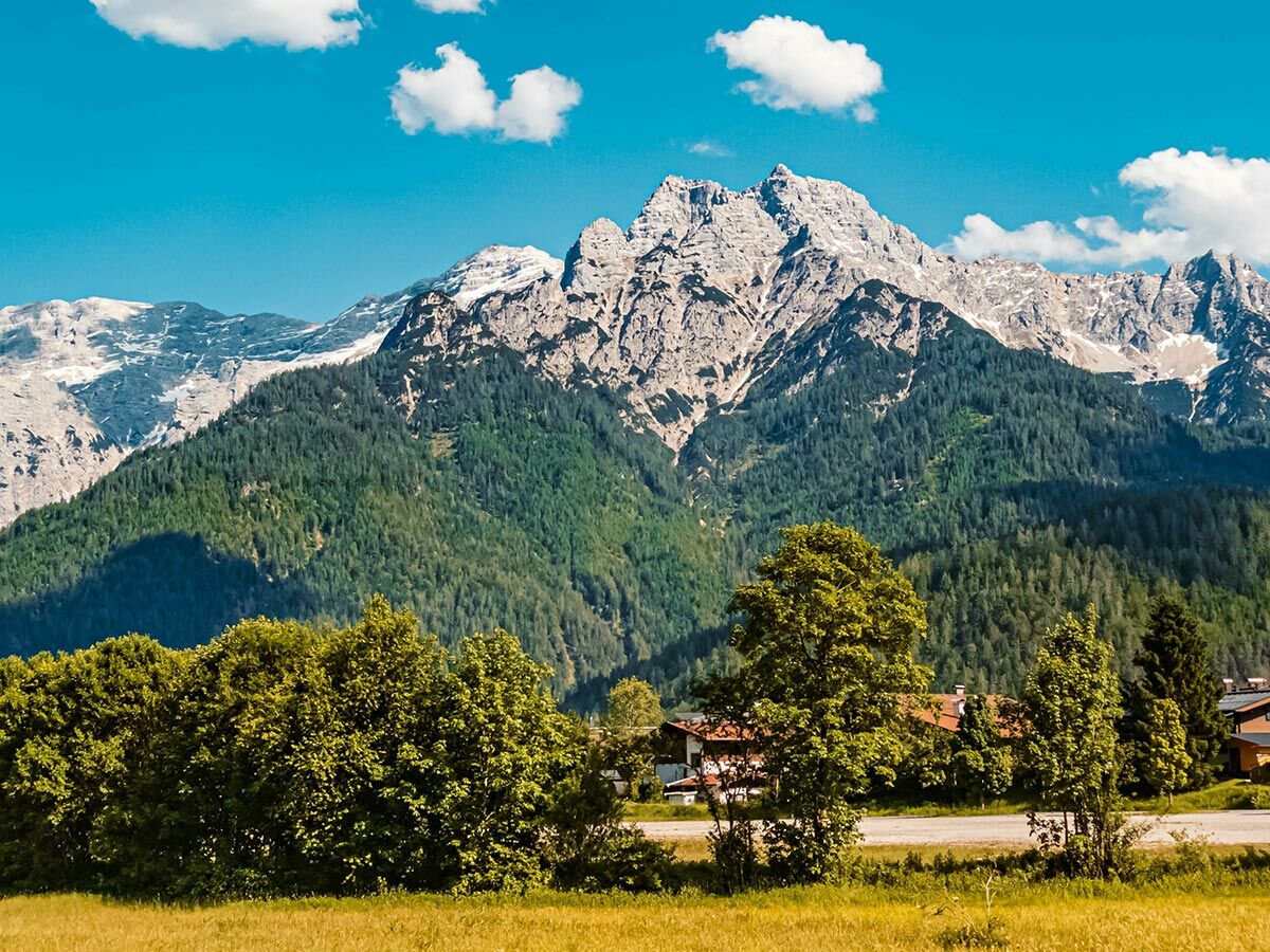 Majestätische Berge mit Schnee, dichte Wälder, sonnige Wiesen und Häuser im Tal unter blauem Himmel mit Wolken.