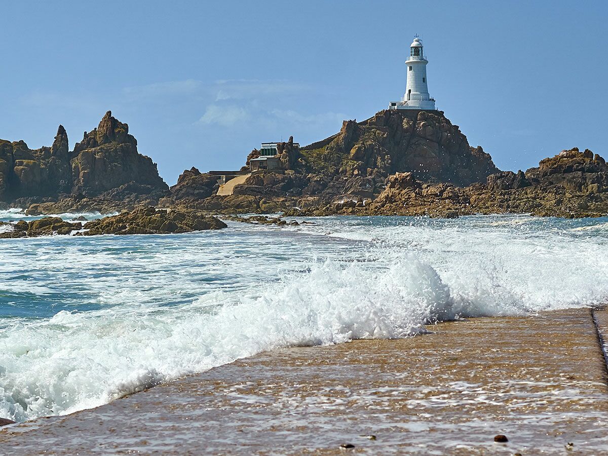 Corbière Leuchtturm auf felsiger Insel, blaue See, Wellen brechen an Damm und Felsen.