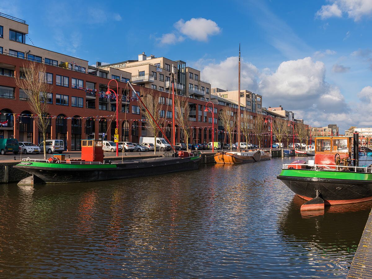 Blick auf einen städtischen Kanal mit vielen geparkten Booten, modernen Gebäuden und Autos entlang des Ufers bei Sonnenschein.