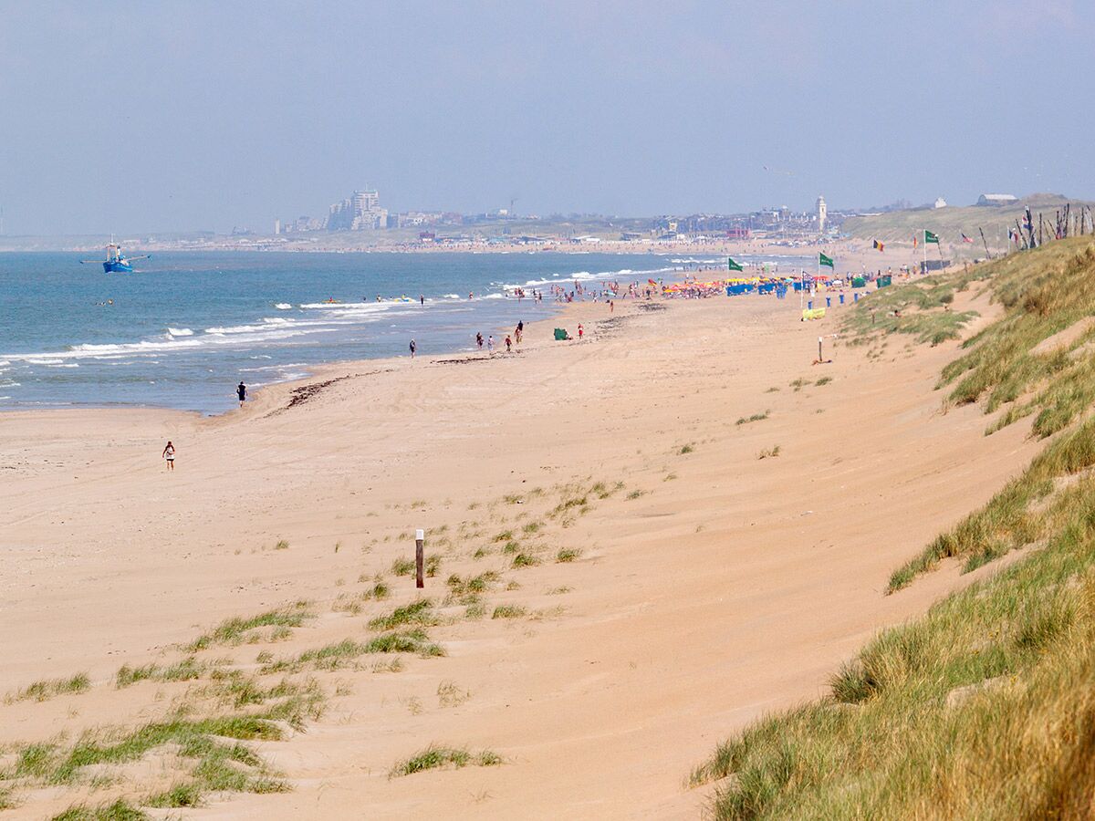 Panoramablick auf einen belebten Sandstrand mit Meer, Wellen, vielen Menschen, Dünen und einer Stadt im Hintergrund.