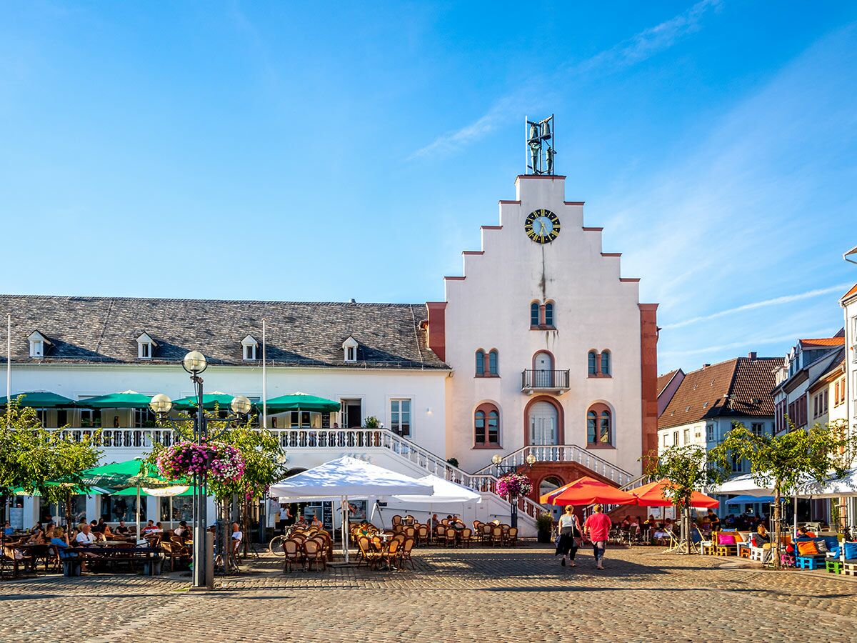 Lebhafter Marktplatz mit historischem Rathaus, Menschen genießen sonnigen Sommertag in Straßencafés unter grünen Sonnenschirmen.