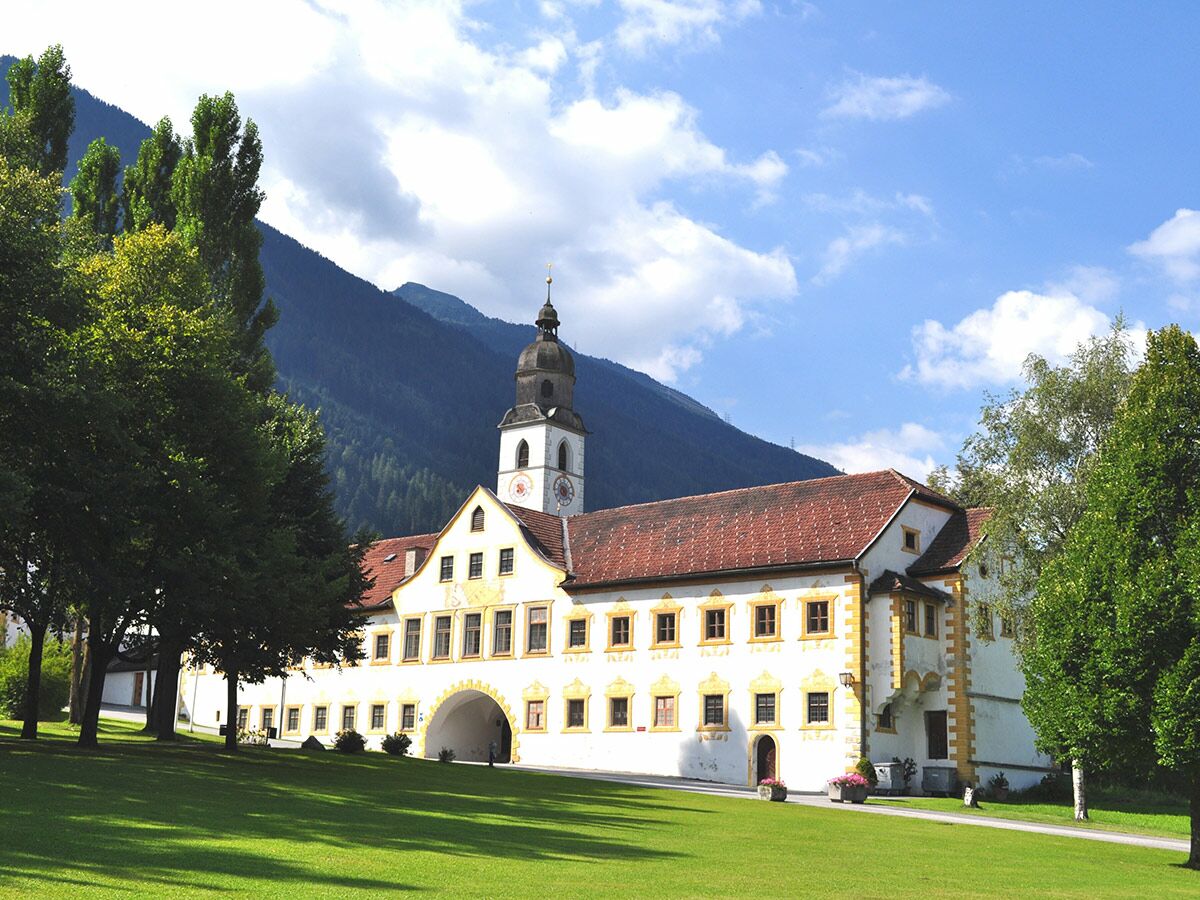 Historisches Benediktinerkloster mit Glockenturm, rotem Dach und gelben Verzierungen vor alpiner Berglandschaft und grünem Rasen.