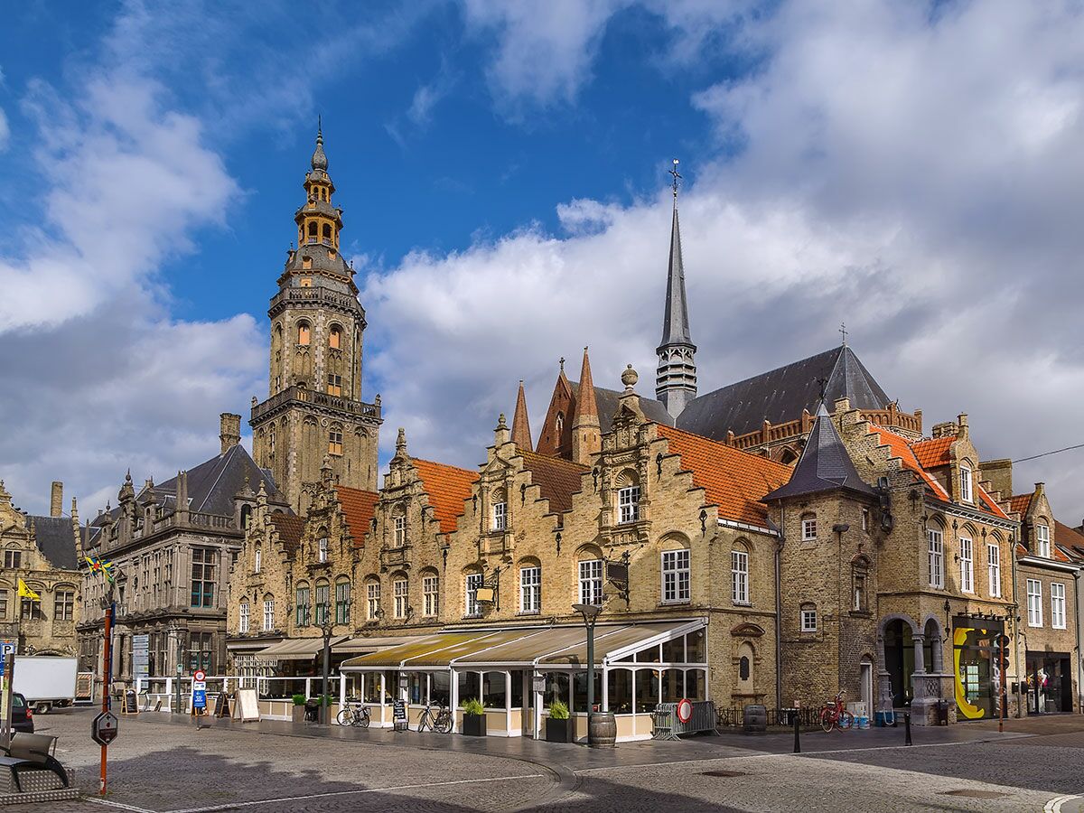 Blick über den Marktplatz von Diksmuide, Belgien, mit historischen Stufengiebelhäusern, einem imposanten Glockenturm und einer Kirchturmspitze unter blauem Himmel.
