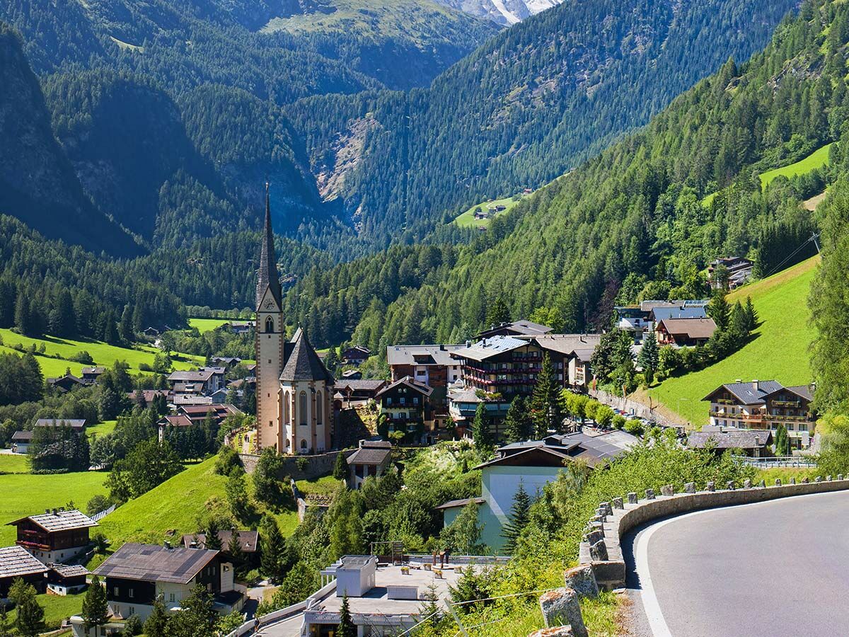 Idyllisches Alpendorf mit hoher Kirche, grünen Wiesen und bewaldeten Bergen unter blauem Himmel an sonnigem Sommertag. Kurvenreiche Straße im Vordergrund.