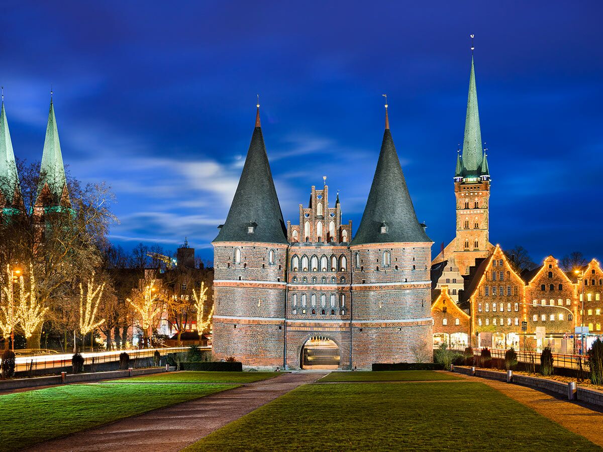 Das historische Holstentor in Lübeck bei Nacht mit blauer Stunde, festlicher Beleuchtung und angrenzenden Gebäuden.