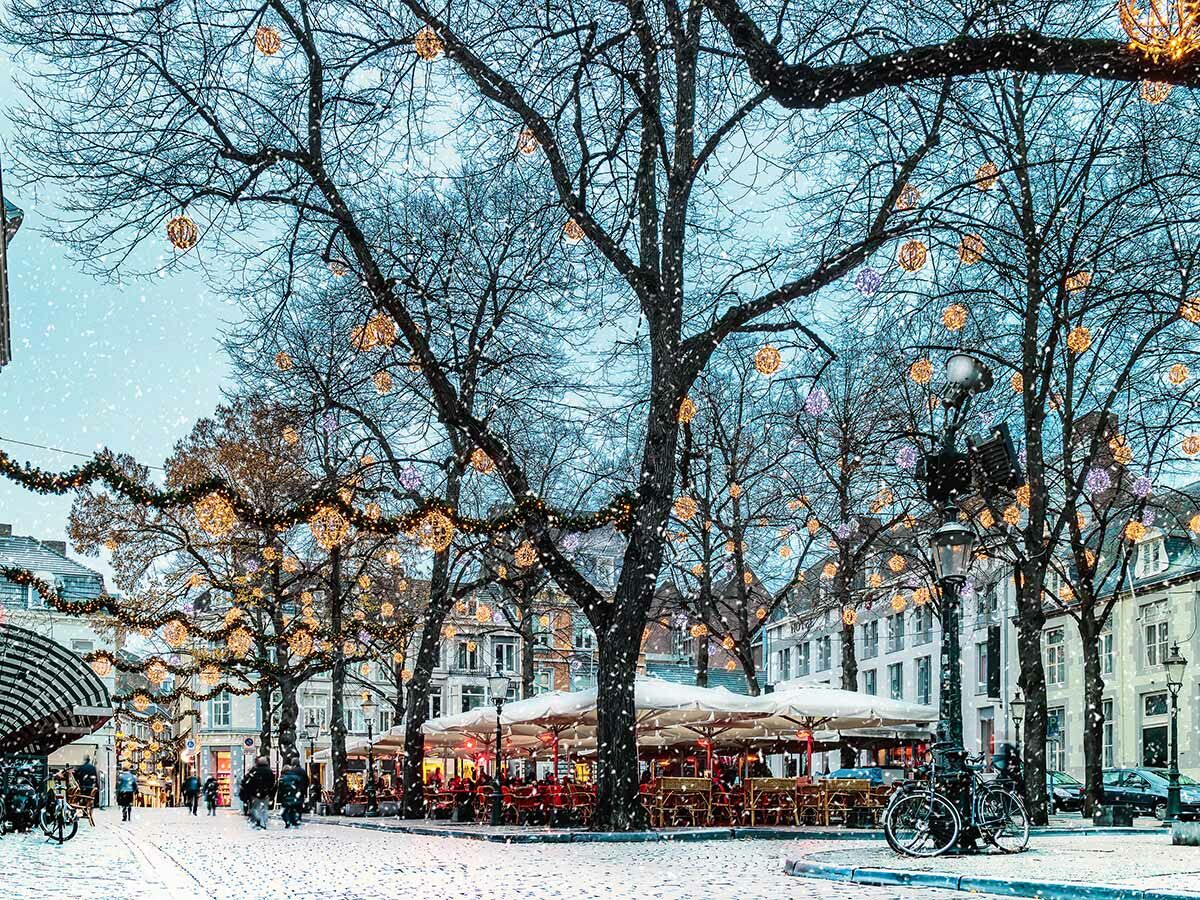 Verschneite winterliche Stadtstraße mit festlicher Weihnachtsbeleuchtung an kahlen Bäumen und Gebäuden, Menschen laufen, Cafés mit Außensitzplätzen.