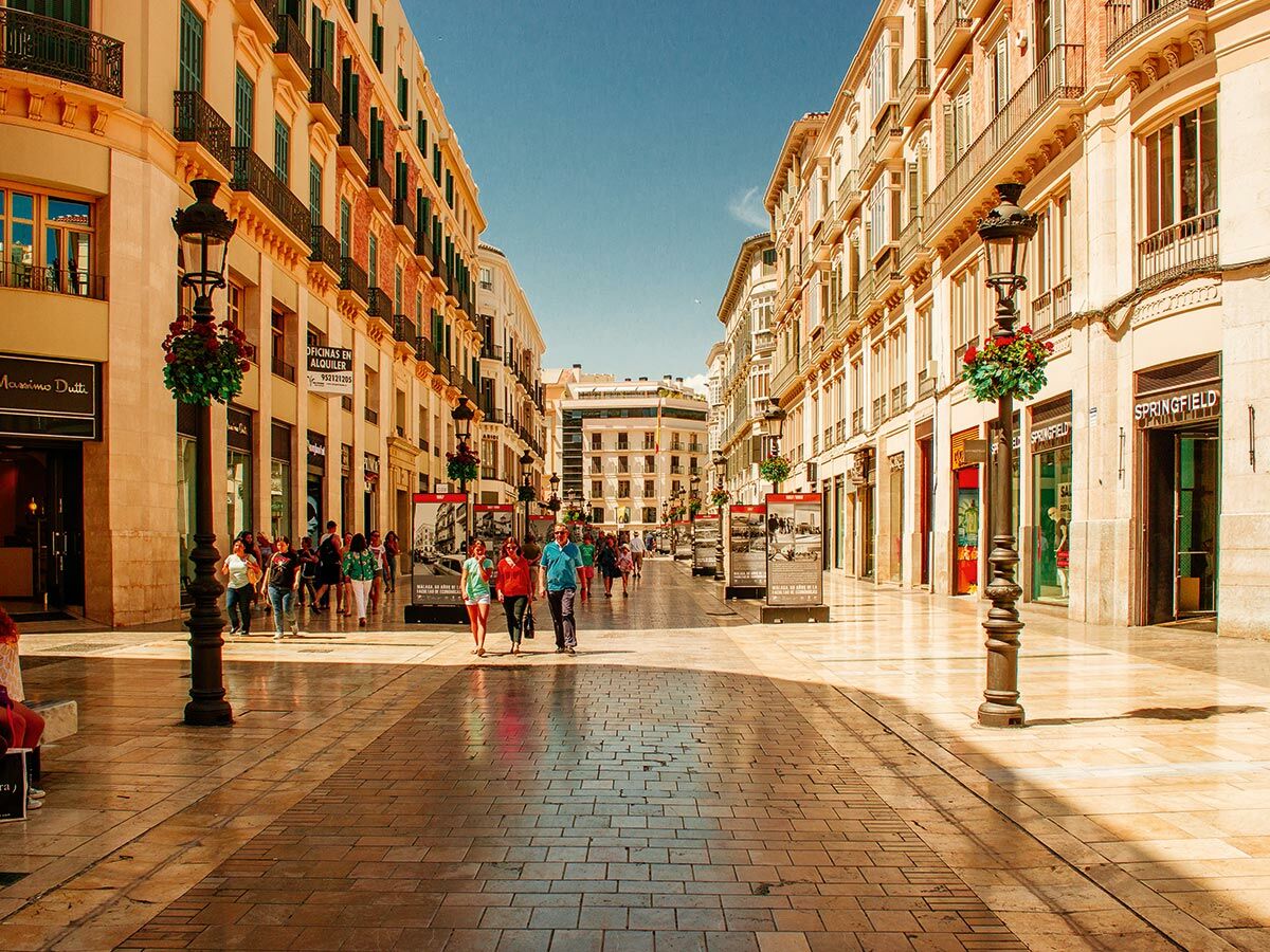 Menschen spazieren auf einer sonnigen Fußgänger-Einkaufsstraße in Málaga, gesäumt von historischen Gebäuden und Geschäften.