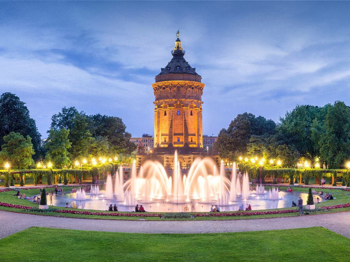 Der beleuchtete Mannheimer Wasserturm im Luisenpark bei Abenddämmerung, umgeben von einem großen Brunnen und entspannten Besuchern.
