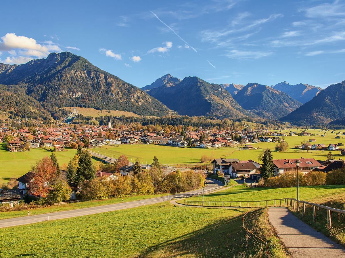 Malerisches Alpendorf im Herbsttal, umgeben von grünen Wiesen, bewaldeten Bergen und einer Skisprungschanze.