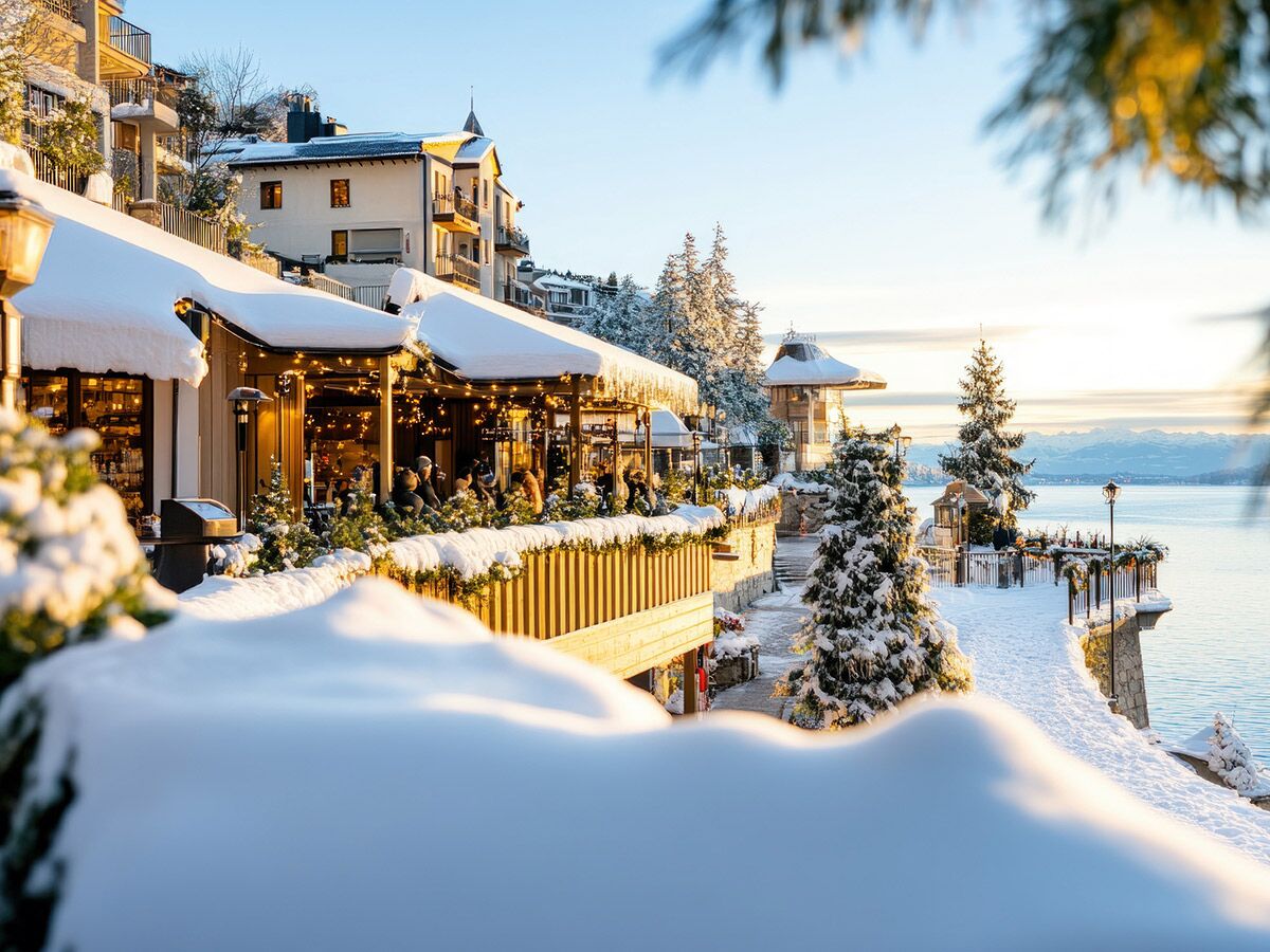 Verschneites Restaurant am See mit festlicher Beleuchtung, winterlicher Atmosphäre und schneebedeckten Bergen im Hintergrund.