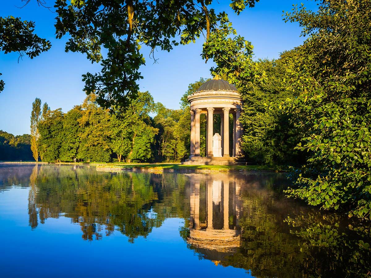 Klassischer Rundtempel mit Statue am ruhigen See, umgeben von grünen Bäumen unter blauem Himmel, mit Spiegelung im Wasser.