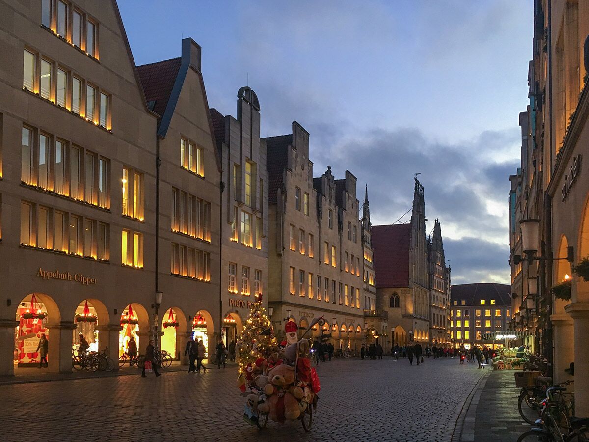 Abendliche Szene in der Altstadt von Münster mit beleuchteten Geschäften und einem festlich geschmückten Wagen mit Teddybären und Weihnachtsbaum.