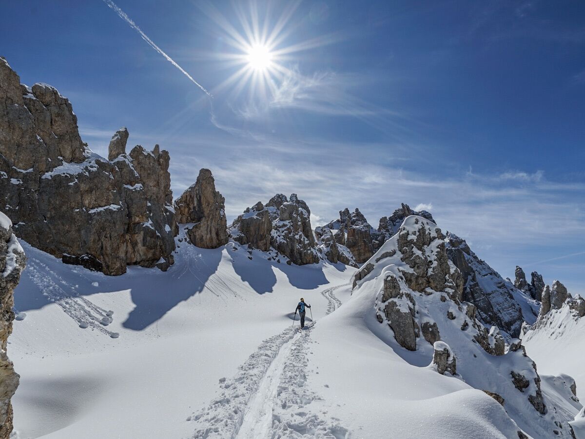 Skitourengeher auf Pfad in verschneiter Gebirgslandschaft mit markanten Felsen, blauem Himmel und strahlender Sonne.