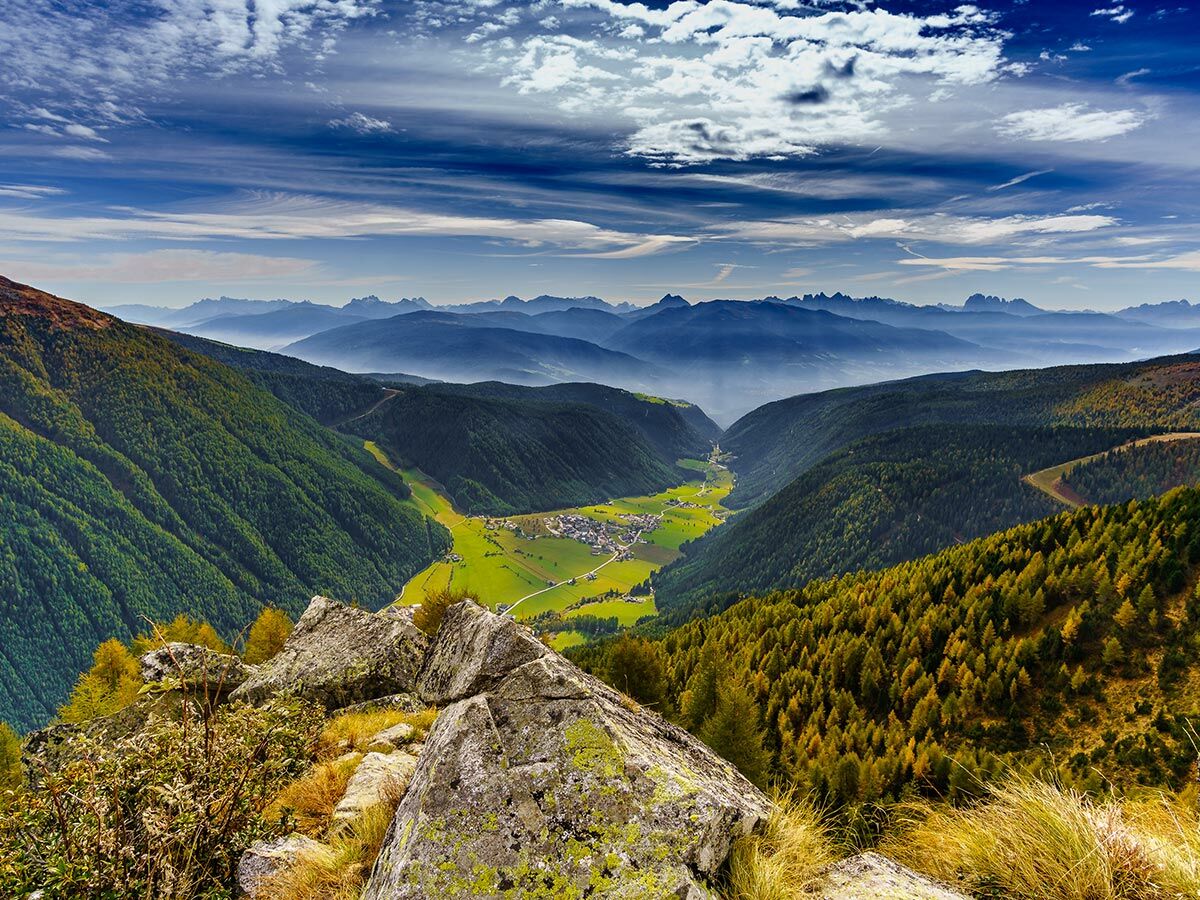 Atemberaubende Aussicht auf ein bewaldetes Bergtal mit einem Dorf in den Dolomiten unter blauem Himmel mit Wolken.