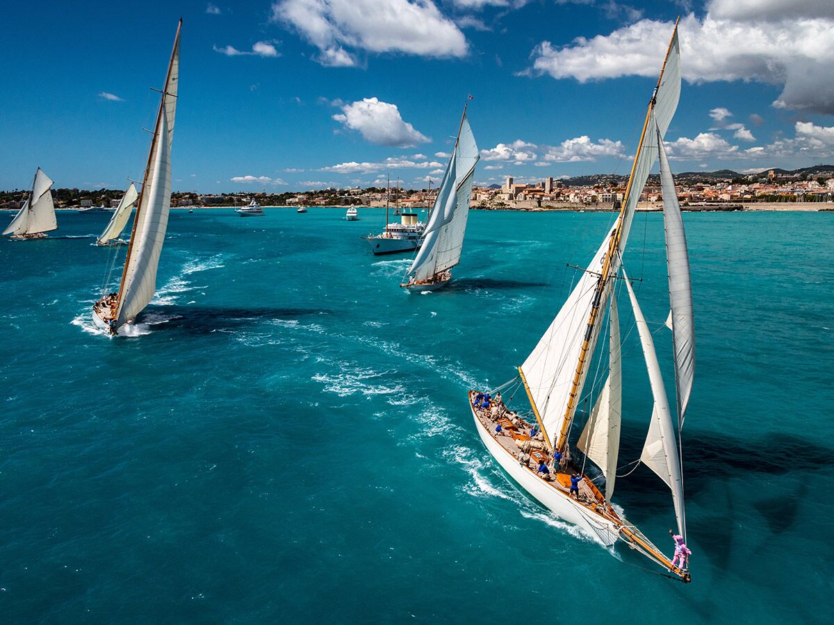 Luftaufnahme klassischer Segelboote, die auf leuchtend türkisfarbenem Wasser nahe einer Küstenstadt unter blauem Himmel segeln.