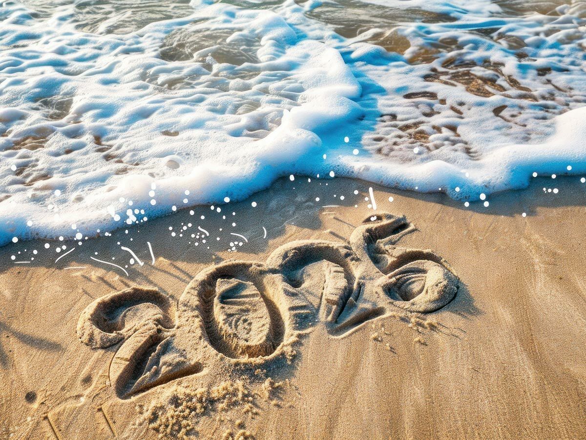 Die Jahreszahl 2026 ist mit Glitzer in den Sand eines sonnigen Strandes geschrieben, während Wellen am Ufer anrollen.