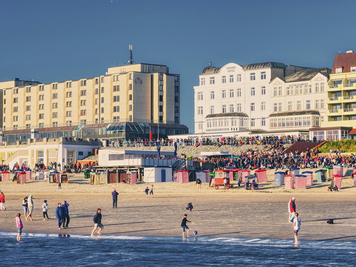 Nordsee-Strand in der Abendsonne mit belebter Promenade, Hotels (Nordsee Hotel, Ostfriesenhof), Strandkörben und spielenden Menschen am Meer.