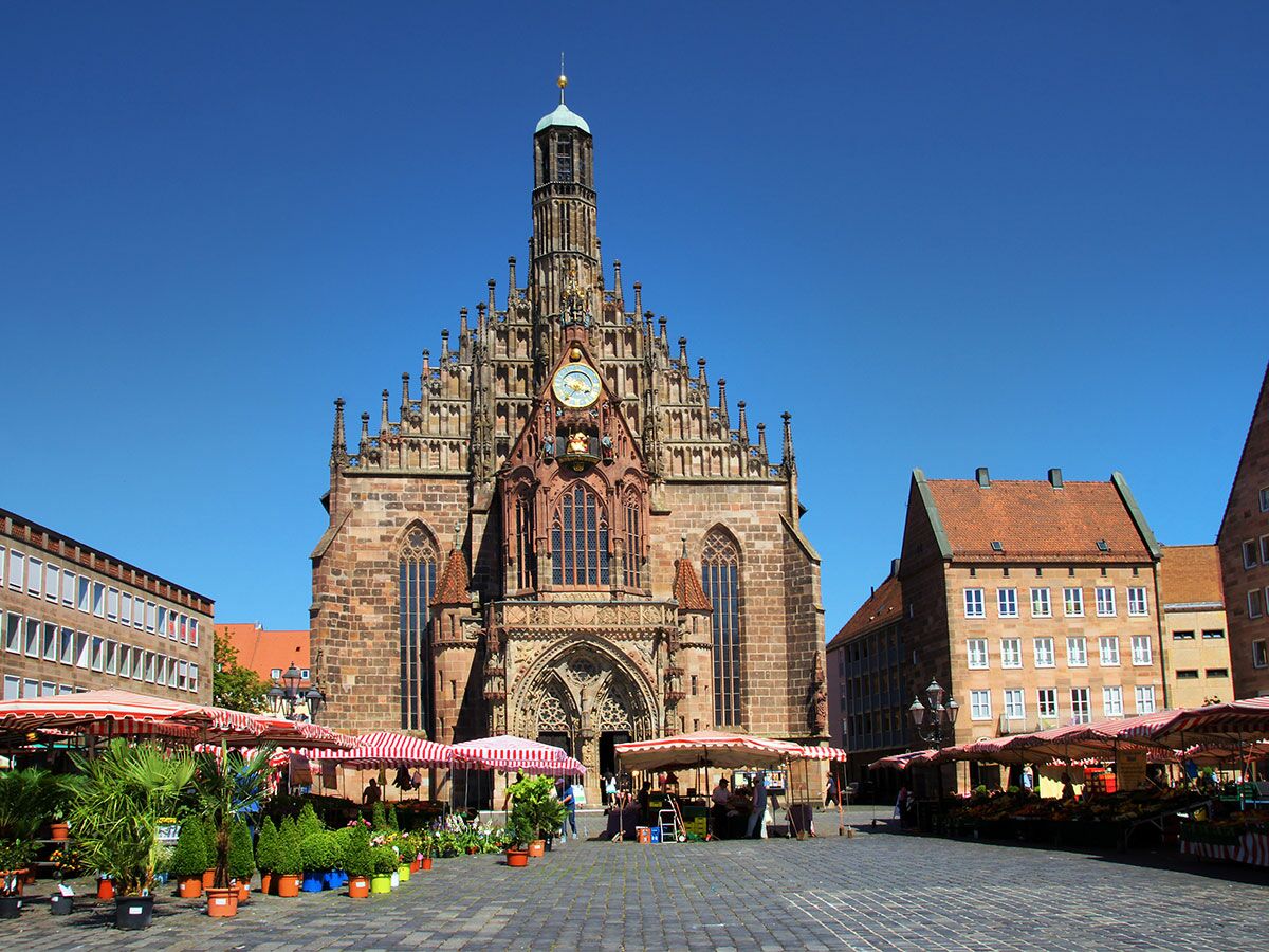 Frauenkirche am Hauptmarkt Nürnberg mit belebtem Markt, roten und weißen Marktständen sowie Pflanzen unter blauem Himmel.