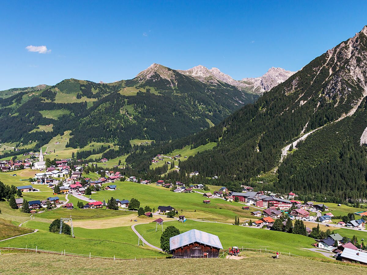 Panoramablick auf ein malerisches Bergdorf in den Alpen mit Kirche, traditionellen Häusern, grünen Wiesen und bewaldeten Bergen unter blauem Himmel.